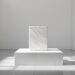 Minimalist white gallery with a marble pedestal and a smaller marble block on top, bathed in sunlight.