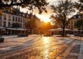 Sunlit city square at sunset with benches, trees, and outdoor café tables; people strolling.