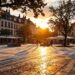 Sunlit city square at sunset with benches, trees, and outdoor café tables; people strolling.