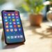 Smartphone resting on a wooden table displaying colorful app icons on the home screen, with a blurred plant and mug in the bright background.