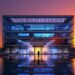 Modern glass-front office building at sunset, with blue-lit interior and a calm reflecting pool showing the façade and a lone figure at the entrance.
